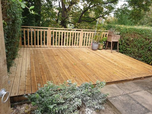 Wooden decking area surrounded by greenery, featuring a wooden railing and a pot plant.