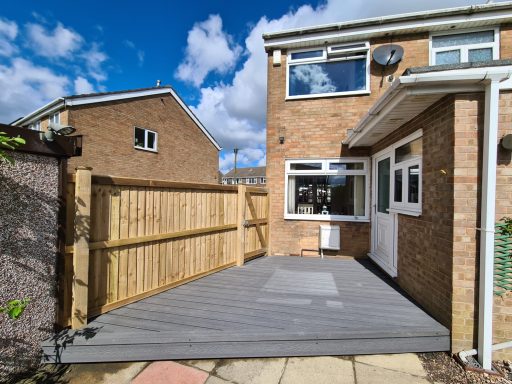 Garden with Grey composite decking with wooden fencing and brick house under a partly cloudy sky.