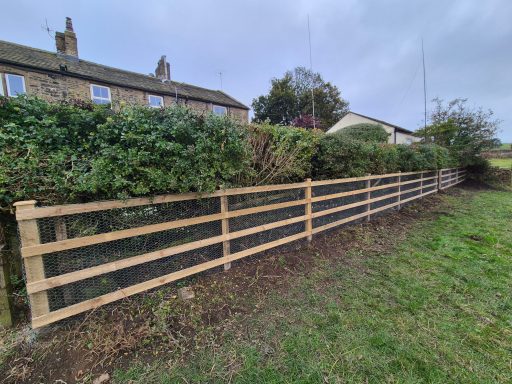 Wooden fence with netting alongside hedges and nearby houses in a rural setting.