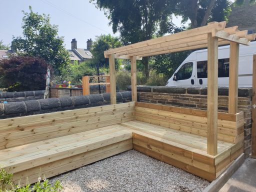 Wooden outdoor seating area with a pergola, surrounded by trees and a gravel surface.