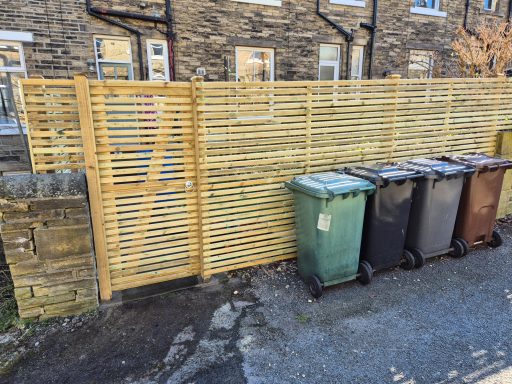 Wooden slatted fence next to three recycling bins in a narrow alleyway.