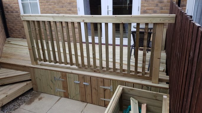 A wooden deck with railings and steps, featuring storage compartments below.