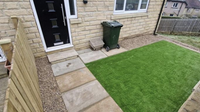 A green artificial lawn beside a stone path and a wheelie bin at a house's side entrance.
