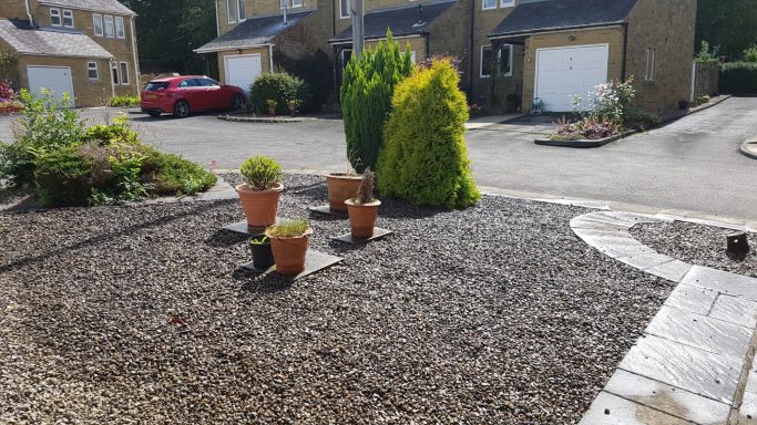 Garden area with potted plants and gravel, near residential houses and a red car.
