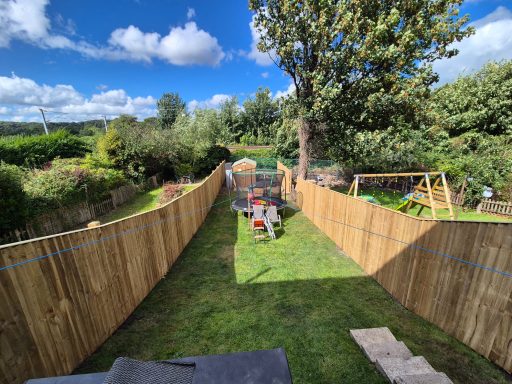 A fenced garden with a grassy area, trees, and outdoor furniture under a blue sky.