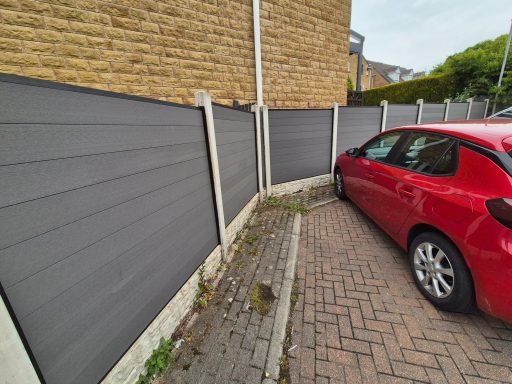 Red car parked closely next to grey fence along a narrow driveway.
